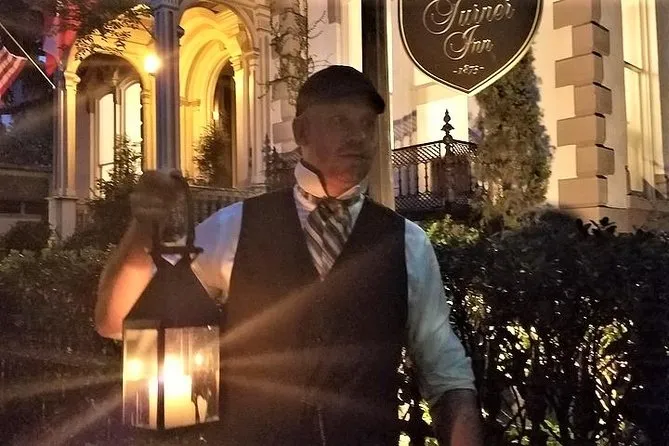 Costumed tour guide in a vest and cap holding a lantern outside a lamplit inn at night