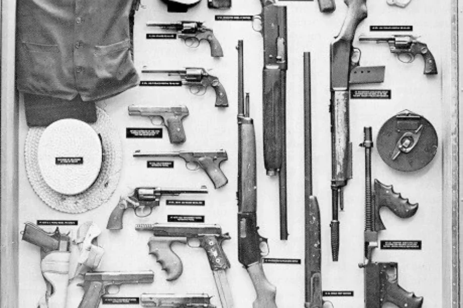 Black-and-white museum case of vintage firearms, a leather jacket, and a straw hat