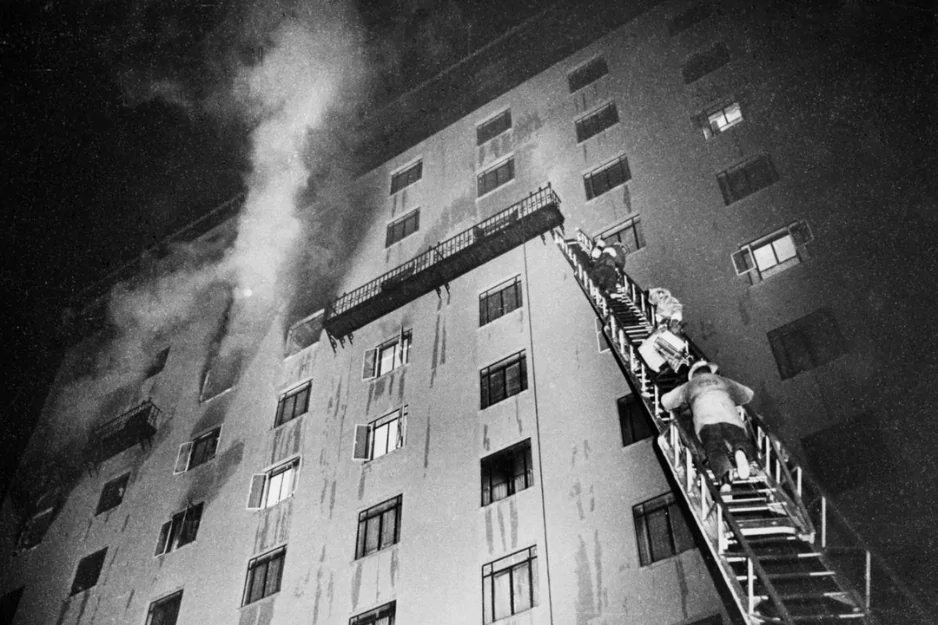 Firefighters climbing a ladder toward a smoking high-rise building at night in black and white