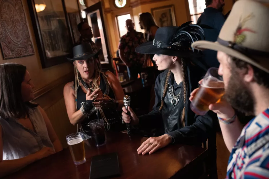 People in feathered hats talking and drinking beer inside a dim historic bar