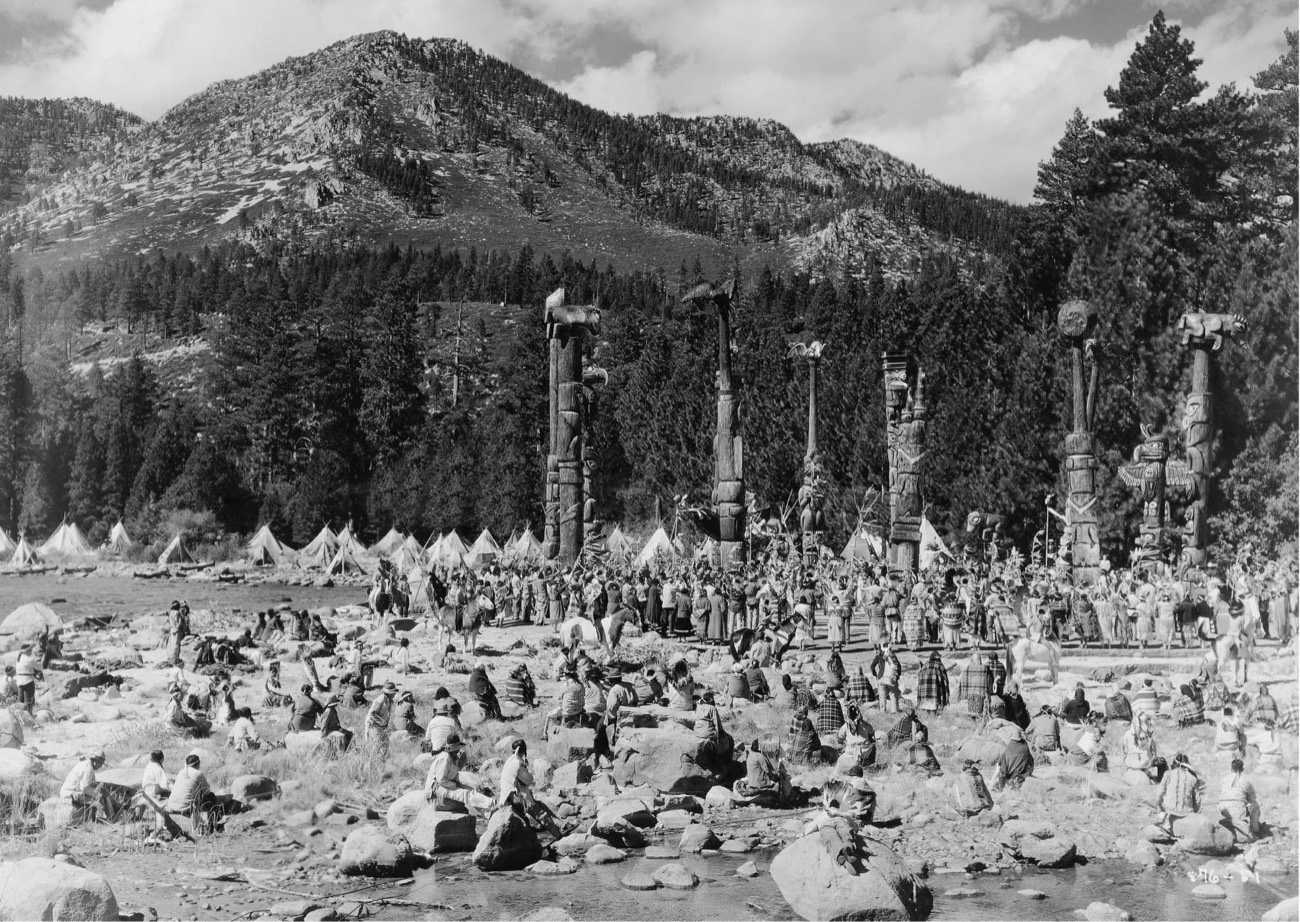 Historic crowd and carved totem poles gathered below a forested mountain