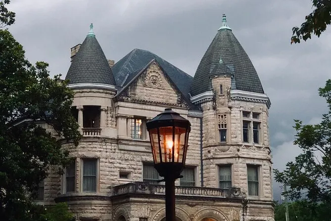 A stone Victorian mansion with turrets behind a lit street lamp at dusk