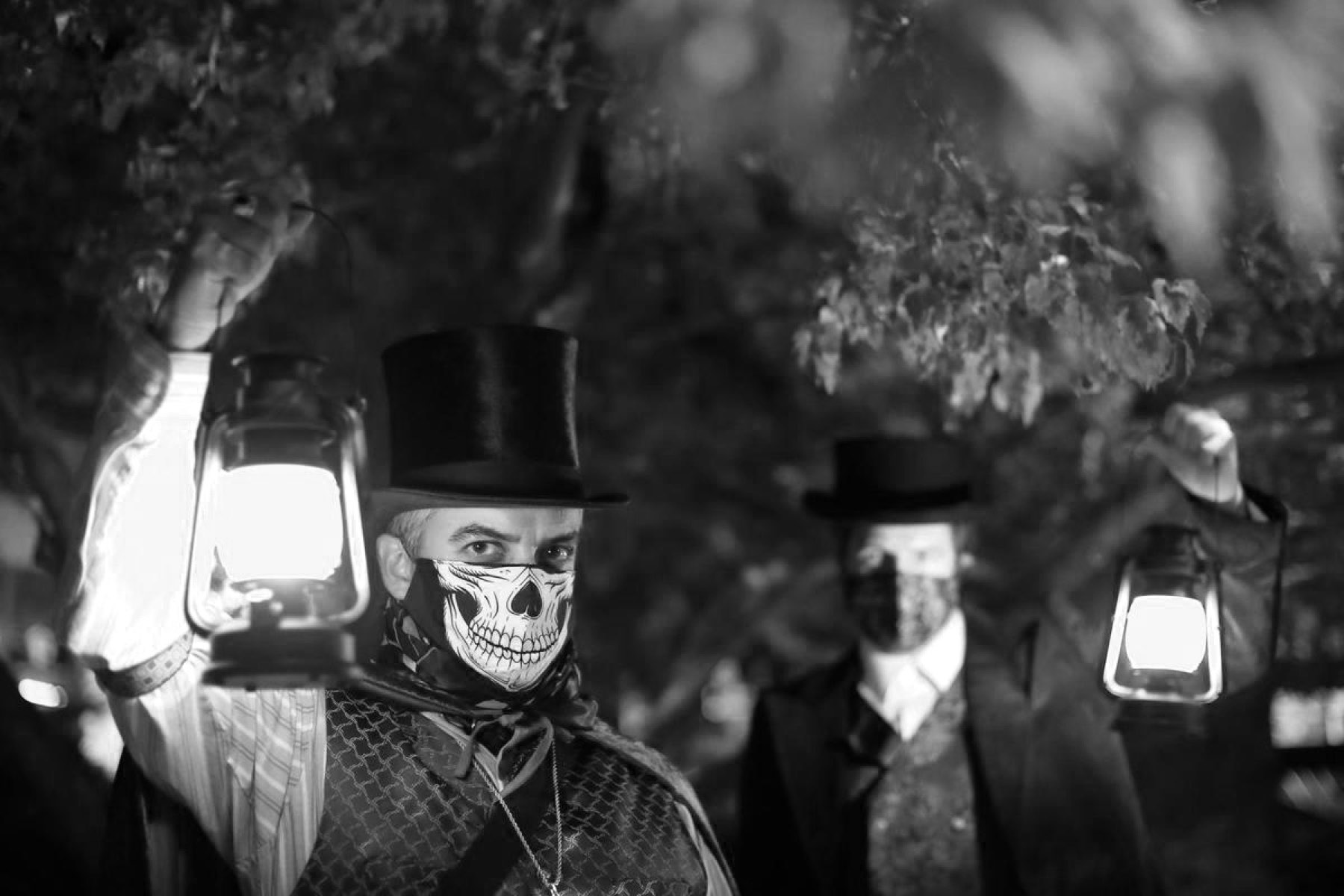 Two costumed guides in top hats and skull masks holding glowing lanterns at night