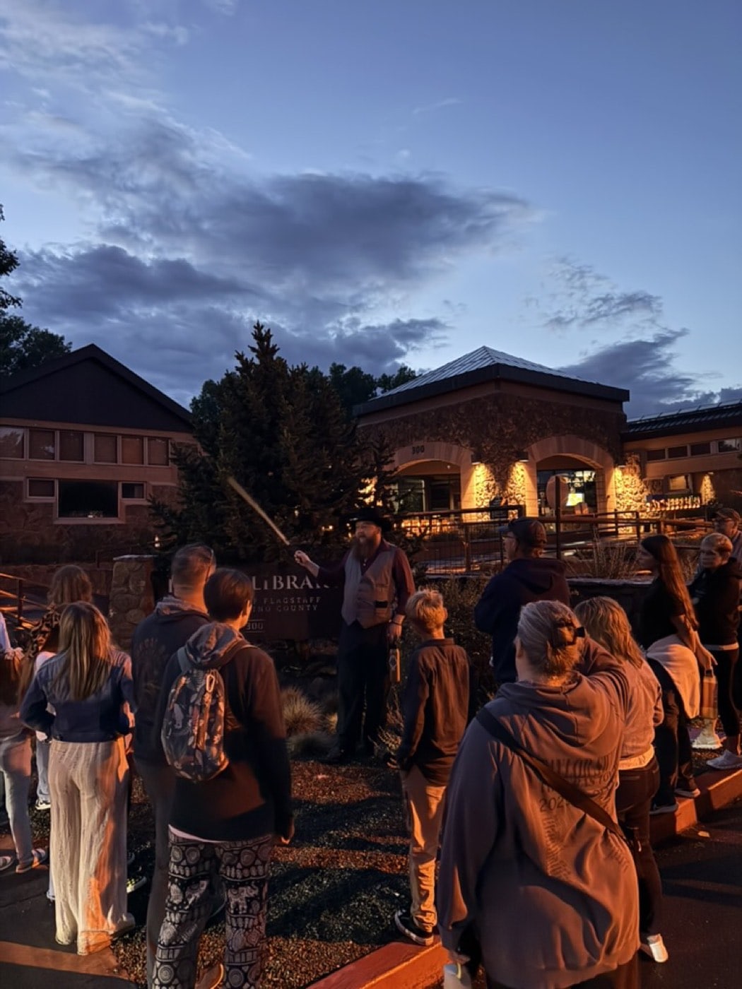 A raised cane punctuates the storytelling as blue hour deepens over the tour group outside the Flagstaff library.