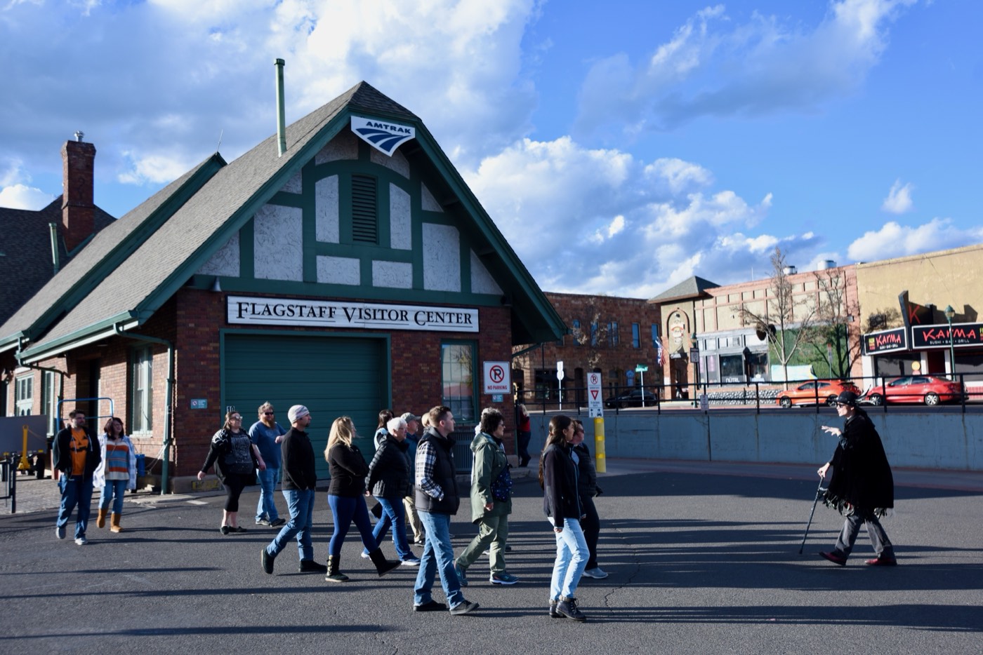 Guests file past the Tudor Revival gable of the Flagstaff Visitor Center, still an Amtrak depot, while their caped guide waits with a cane on the old Route 66 alignment.