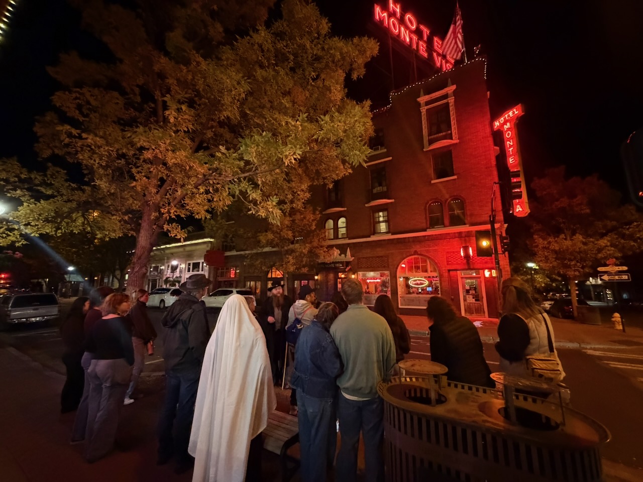 A guest in a white sheet ghost costume stands with the tour group under the red Hotel Monte Vista neon sign in downtown Flagstaff at night