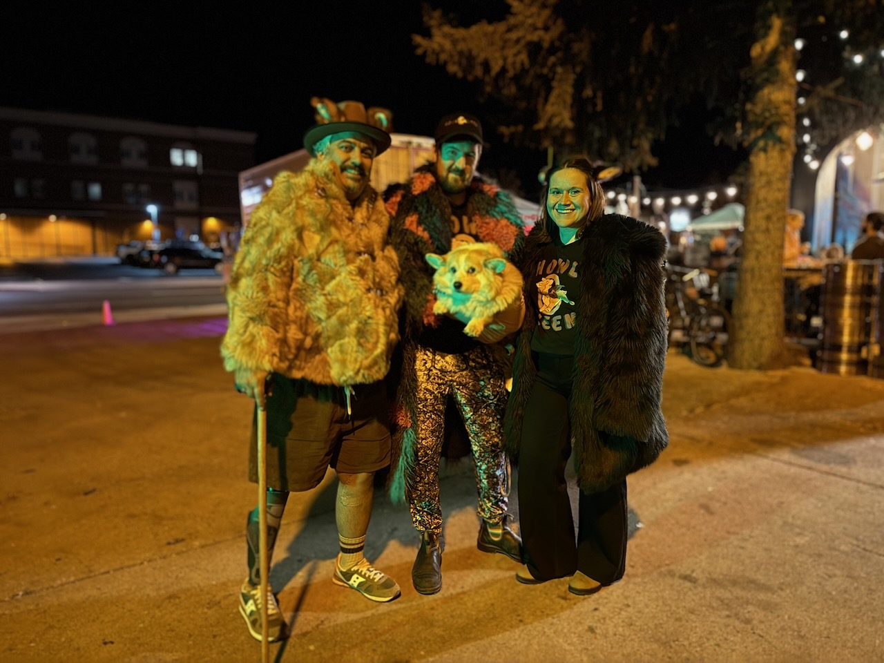 Three people at night at Howl-O-Ween 2025 in downtown Flagstaff, one holding a stuffed corgi