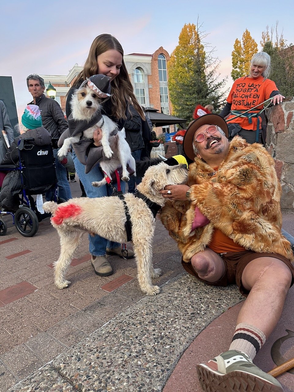 Woman holding a terrier in a shark costume next to a man in a fur coat and a white dog at Howl-O-Ween 2025