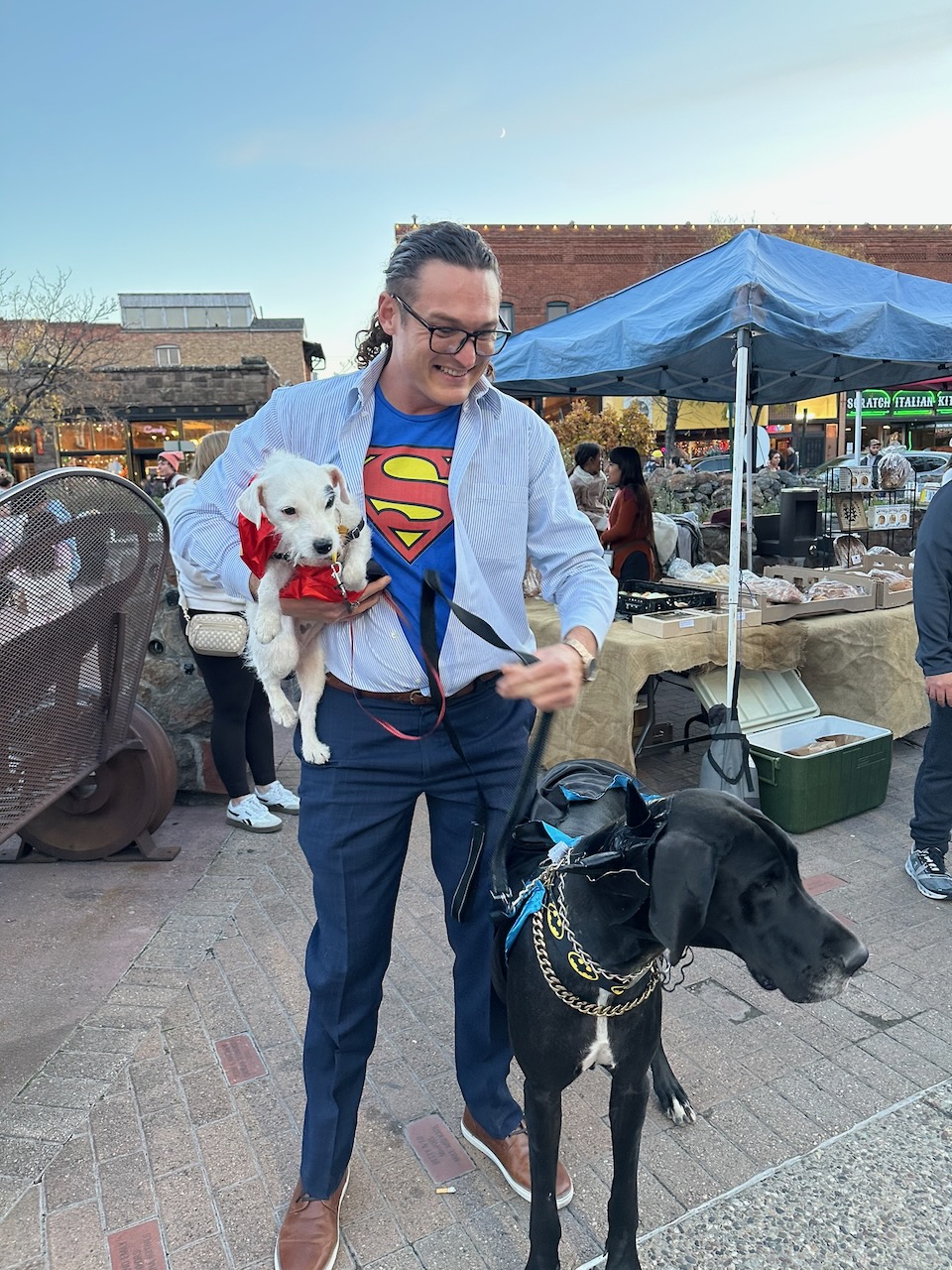 Man in a Superman shirt holding a white puppy and walking a Great Dane at a Howl-O-Ween vendor tent