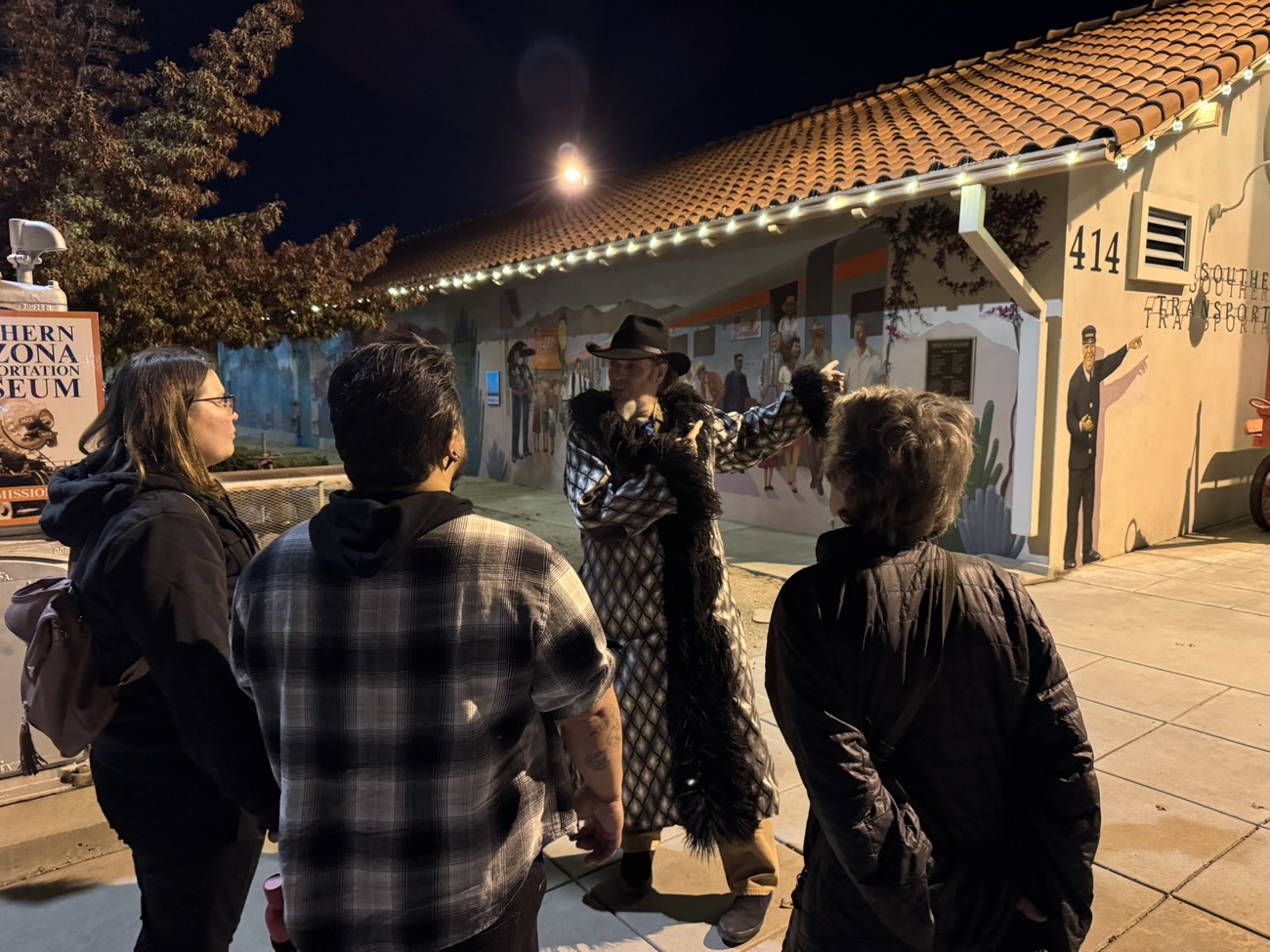 Costumed tour guide addressing a small group at night by the Tucson transportation museum mural